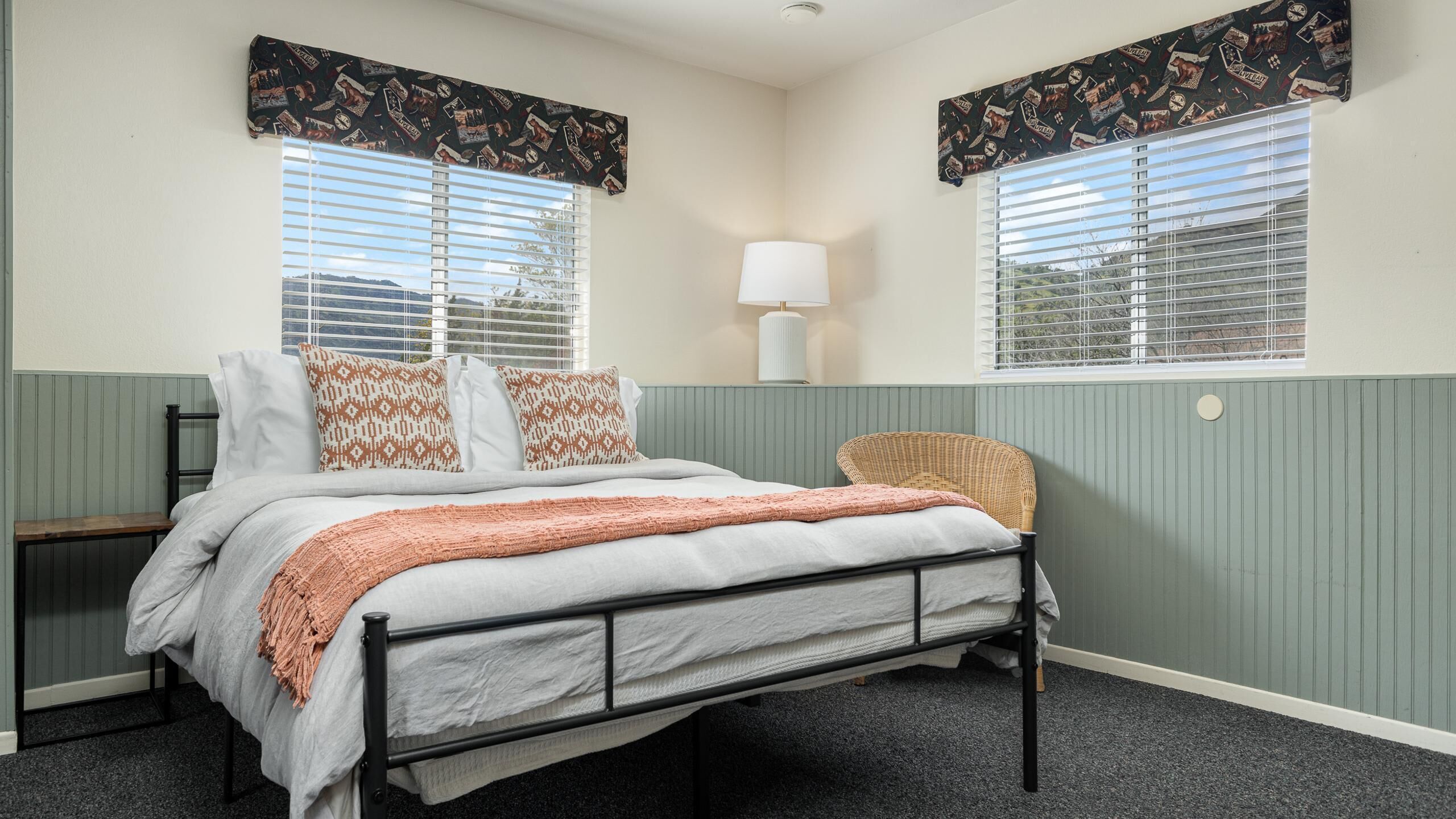 Un dormitorio con cama, mesita de noche, lámpara y dos ventanas con cortinas estampadas.
