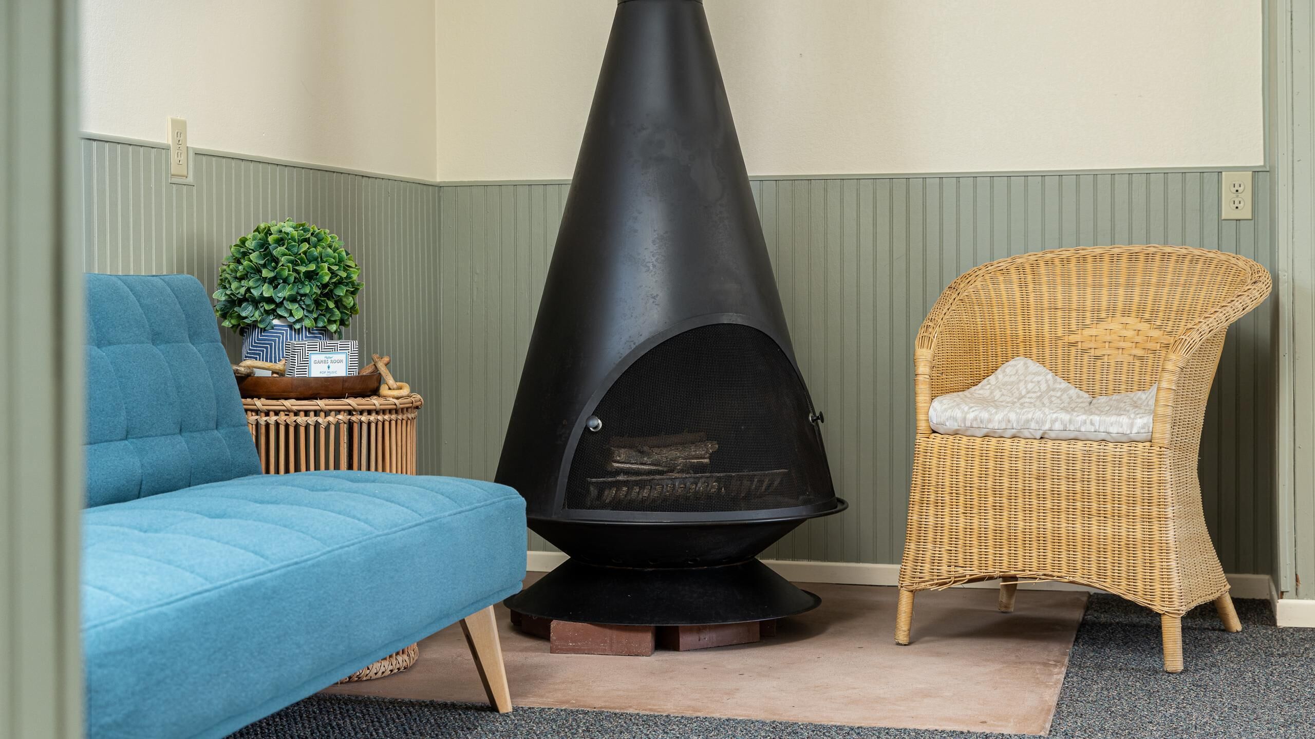 A room with a black wood-burning stove, a blue sofa, and a wicker chair with a cushion.