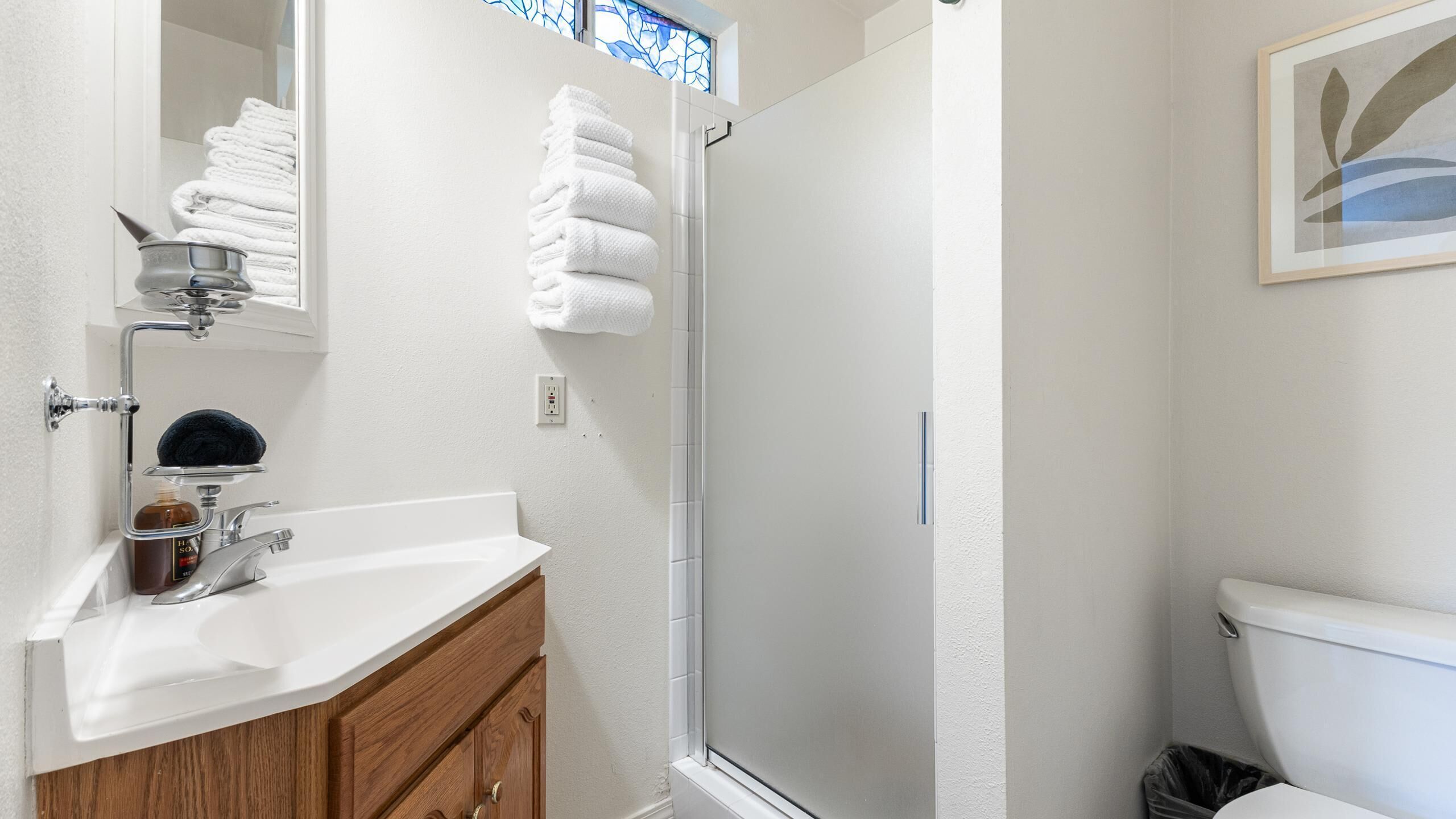 Un baño con lavamanos blanco, mueble de madera y mampara de ducha de vidrio.