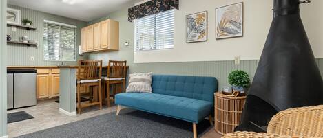 Un espacio habitable compacto con kitchenette, un sofá azul, zona de comedor con sillas de madera y estufa negra.