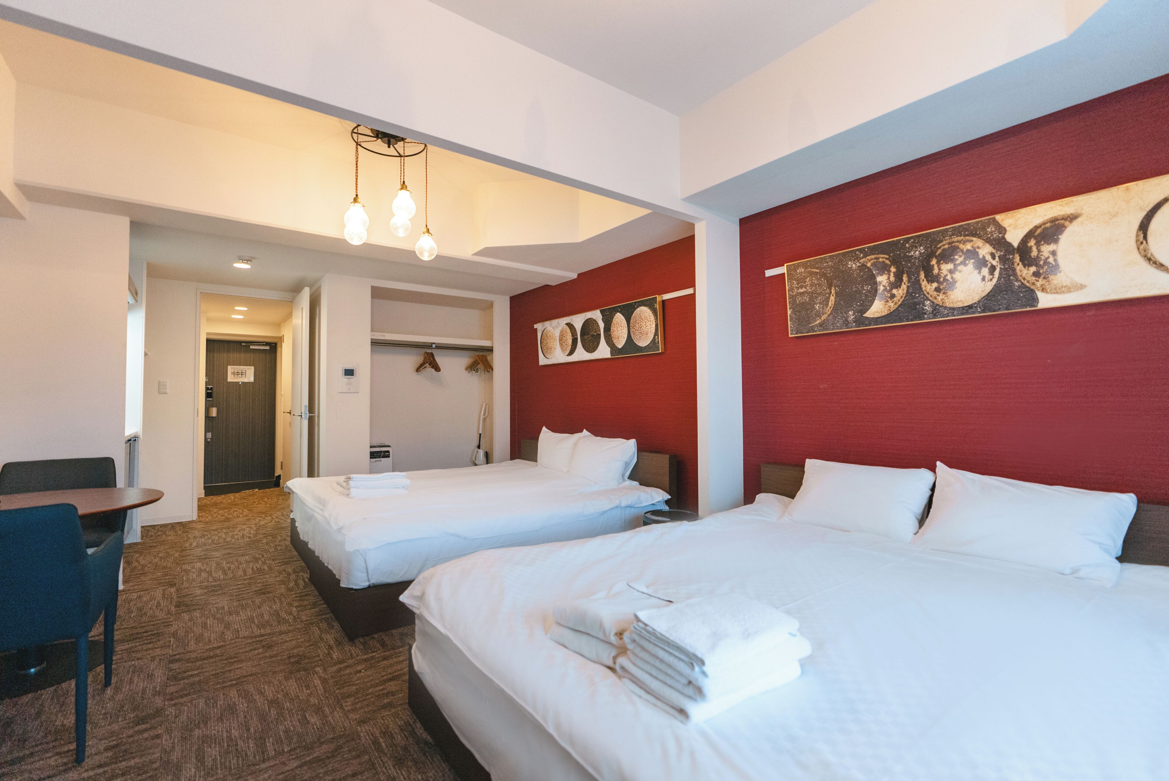 A hotel room with two beds, a desk, and a chair. There is a red accent wall with a moon phase artwork.