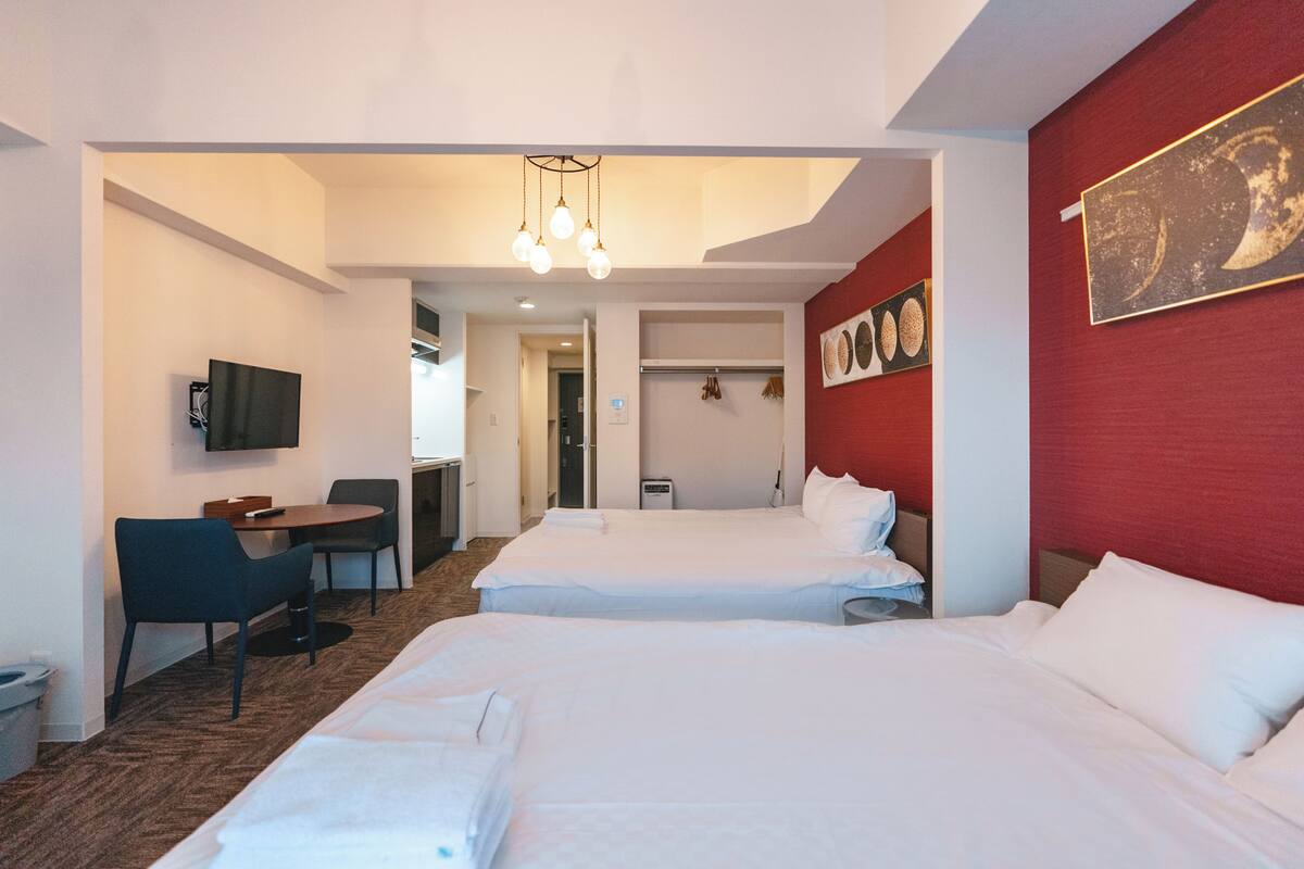 A hotel room with two beds, a desk with a chair, a TV, and a red accent wall with artwork.