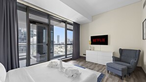 A hotel room with a large bed, a grey armchair, a white dresser, and a TV displaying the Netflix logo.
