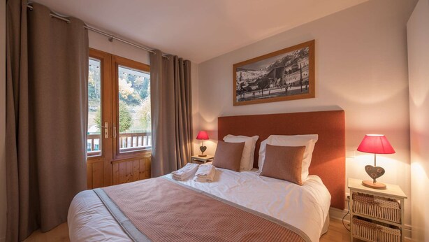 A neatly made bed with a headboard, two bedside lamps, a window with curtains, and a framed picture on the wall. - La Clusaz Primevères summer (La Clusaz)