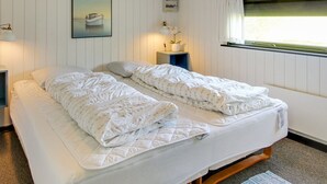 Un dormitorio con cama, mesita de noche, panel eléctrico en la pared, ventana con cortinas e imagen en un marco colgada.