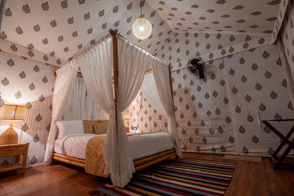 Un lit à baldaquin à quatre poteaux, avec une structure en bois et du linge de lit blanc, un tapis à rayures et un papier peint à motifs représentant des feuilles.