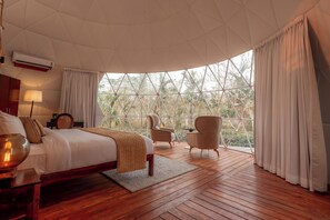 A bedroom with a large bed, a wooden floor, a window with curtains, and a view of trees. - Vaani Greens (Kaliabar)
