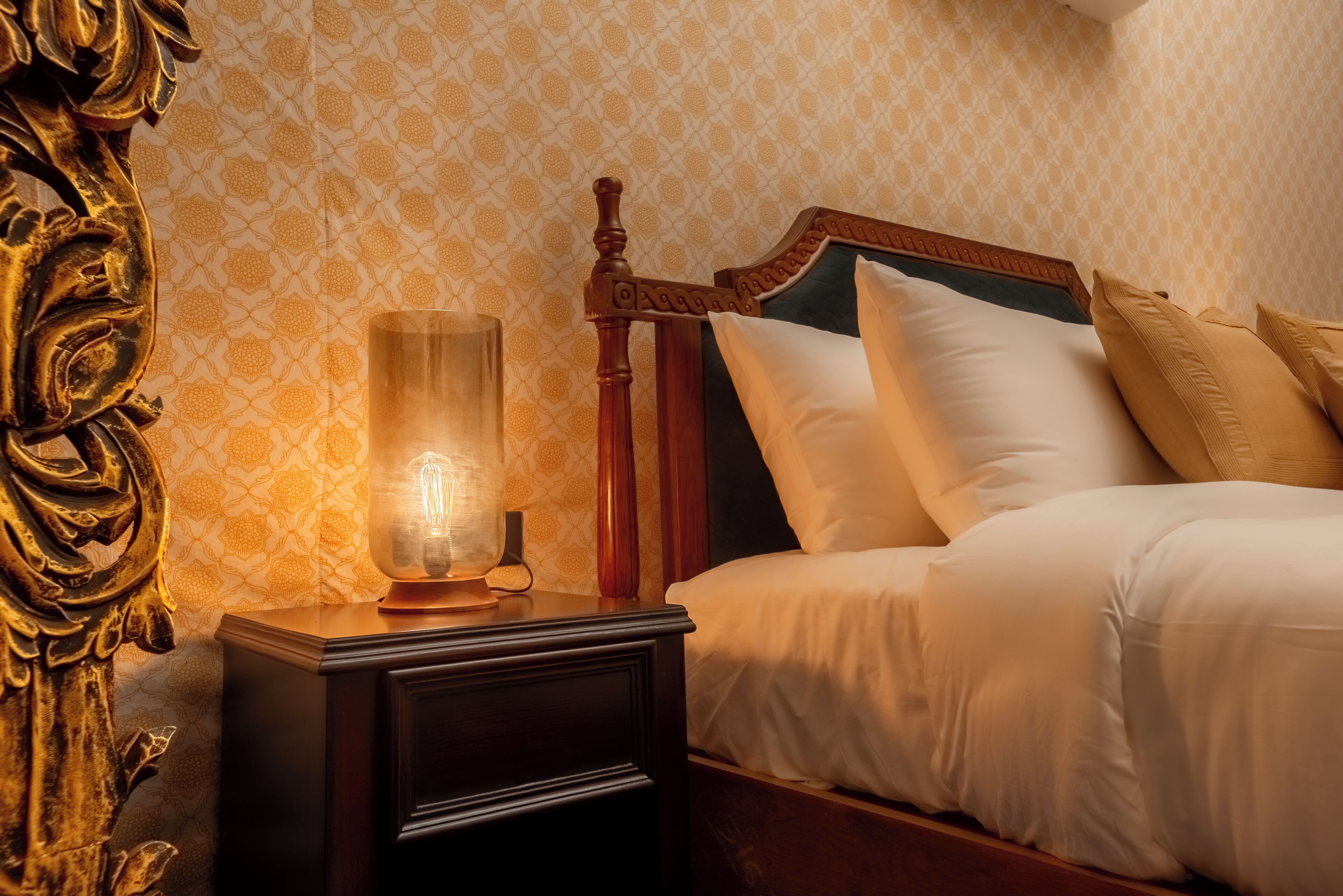A neatly made bed with white linens and a wooden headboard, a bedside table with a lamp, and a wall with a patterned wallpaper.