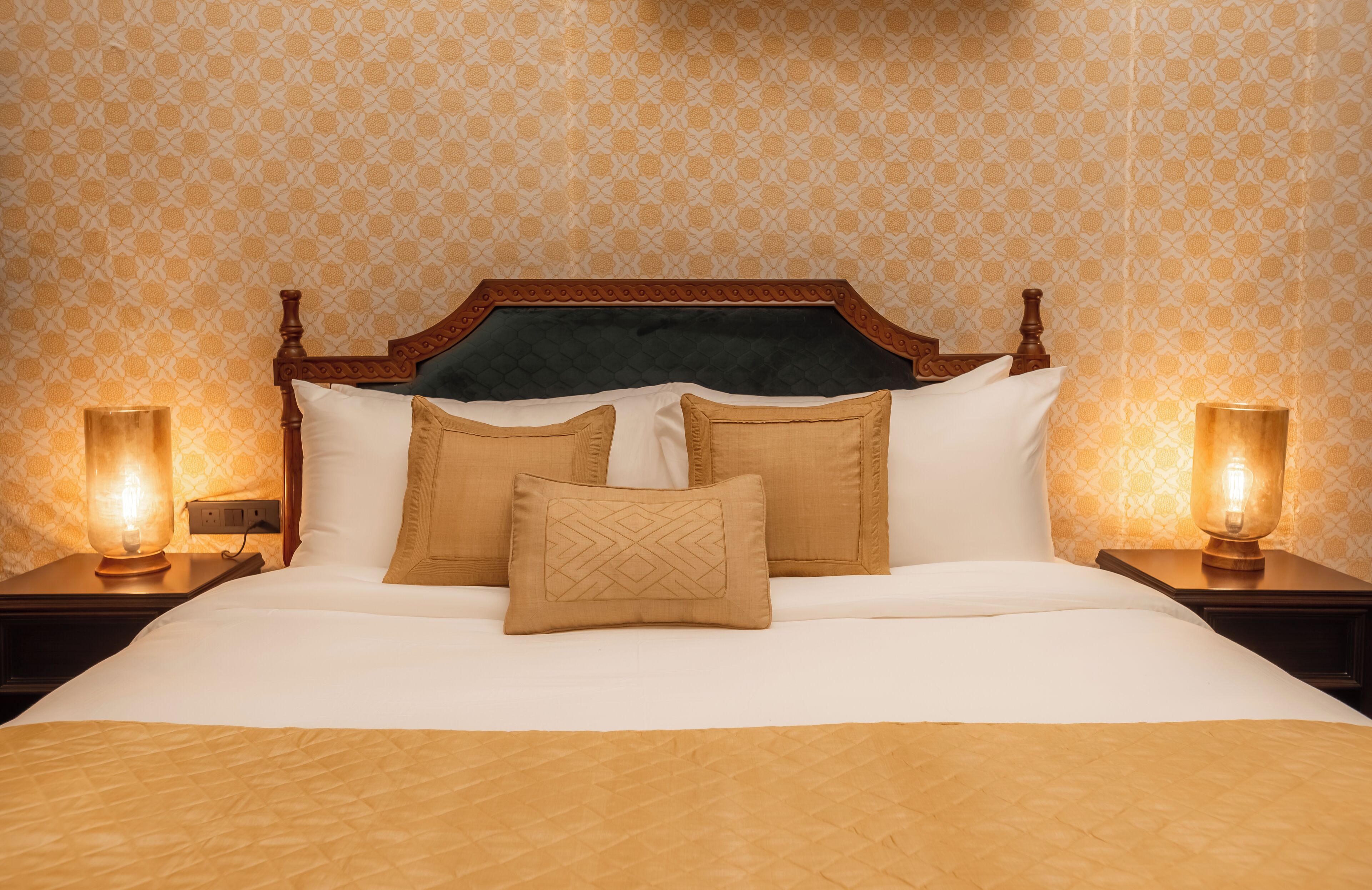A neatly made bed with a wooden headboard, two bedside lamps, and patterned wallpaper.