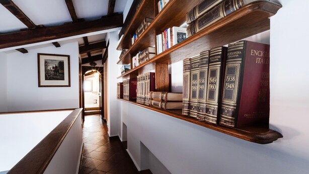 A library with wooden bookshelves filled with books, a framed picture on the wall, and a staircase leading to another room. - Italianway - Bigli 6 (Milan)