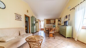 A living room with a sofa, a round wicker table, and a kitchen in the background. - Sa Pedrera in Sa Rocca Tunda (San Vero Milis)