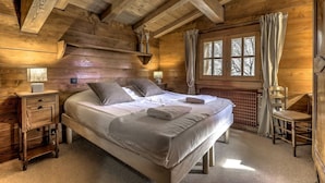 Quarto de cabana de madeira com uma cama, mesas de cabeceira, uma cadeira e uma janela com cortinas.