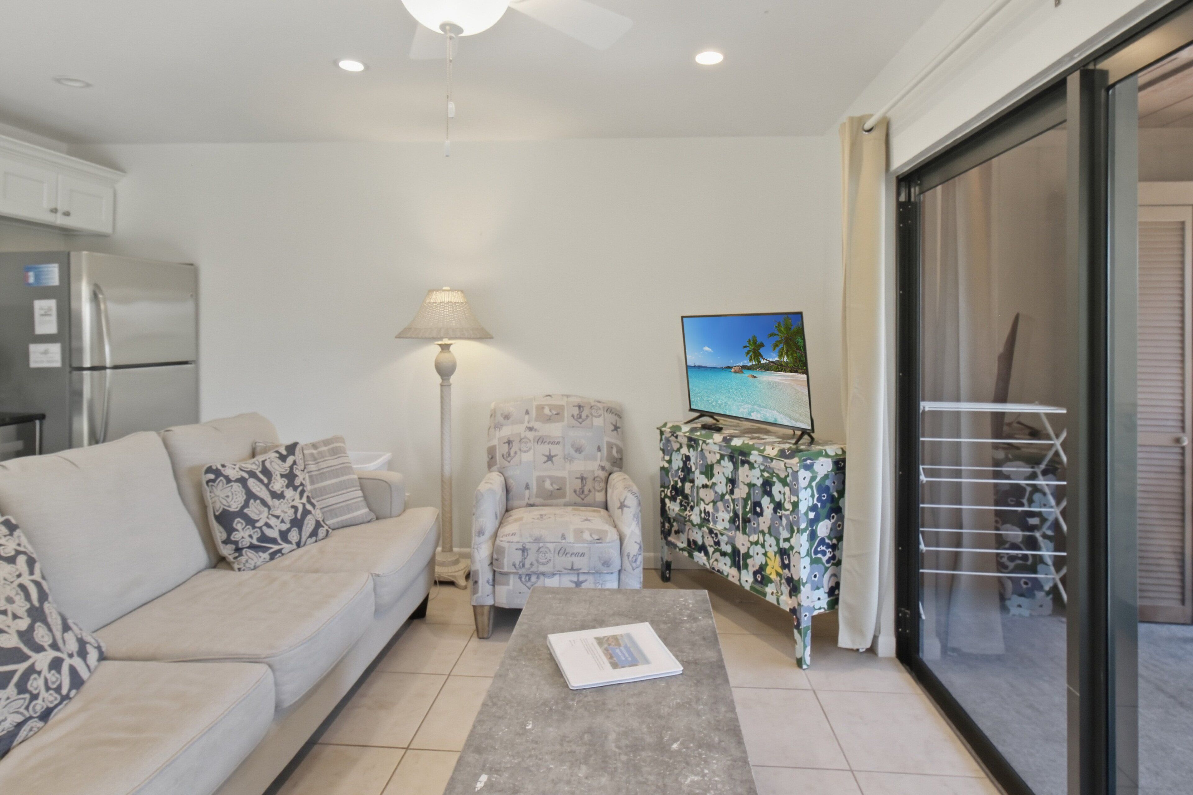 A living room with a sofa, armchair, coffee table, lamp, TV, and a view of the sea through a sliding door.