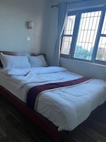 Una cama bien hecha con sábanas blancas y un edredón morado, una lámpara de pared y una ventana con vistas a edificios.