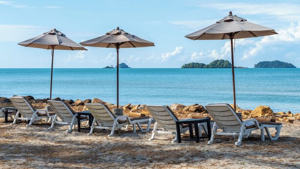 Une plage bordée de rangées de chaises longues et de parasols, un rivage rocheux et une mer calme avec des îles à l’horizon.