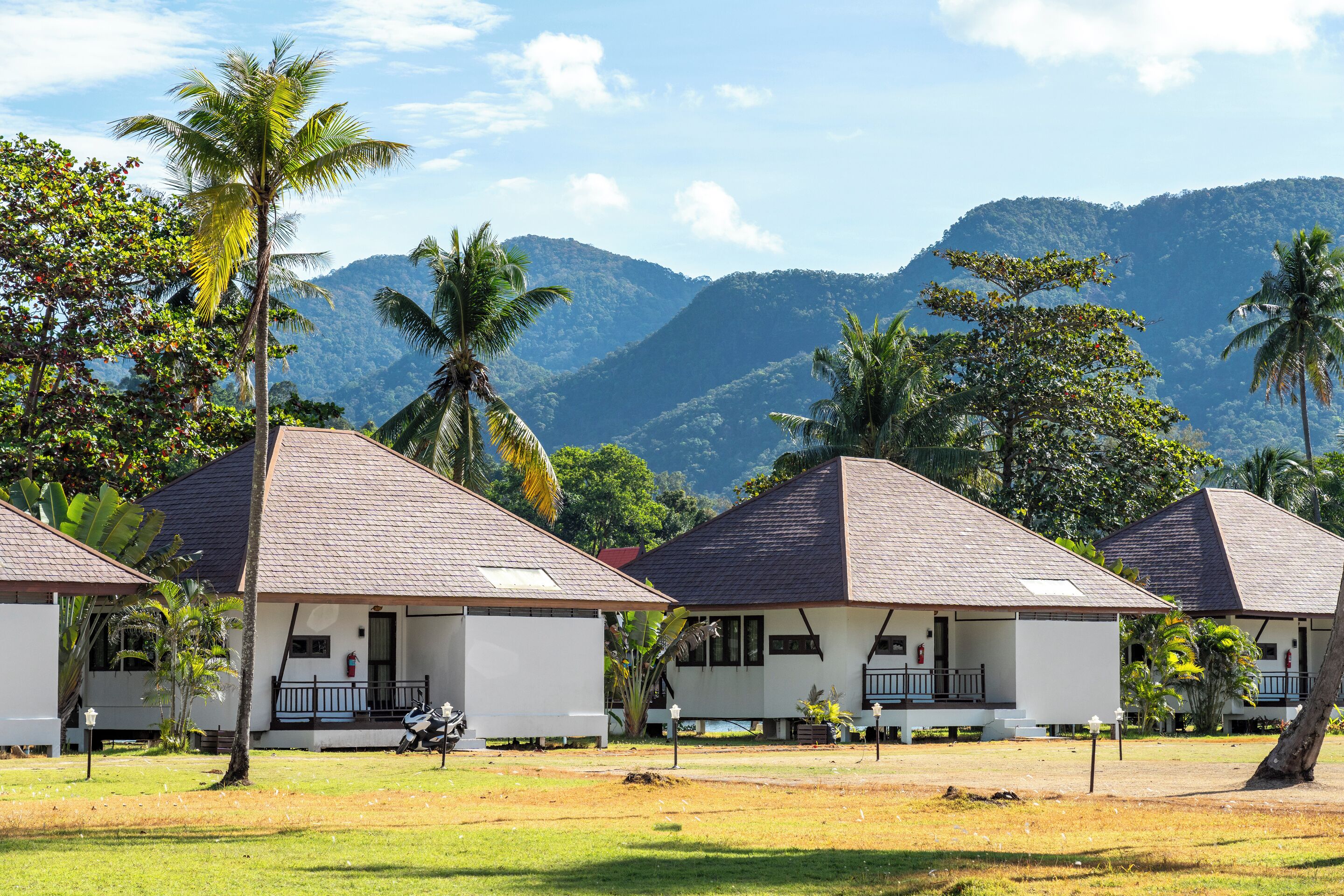 A row of white bungalows with dark roofs, surrounded by lush greenery and palm trees, set against a backdrop of mountains.