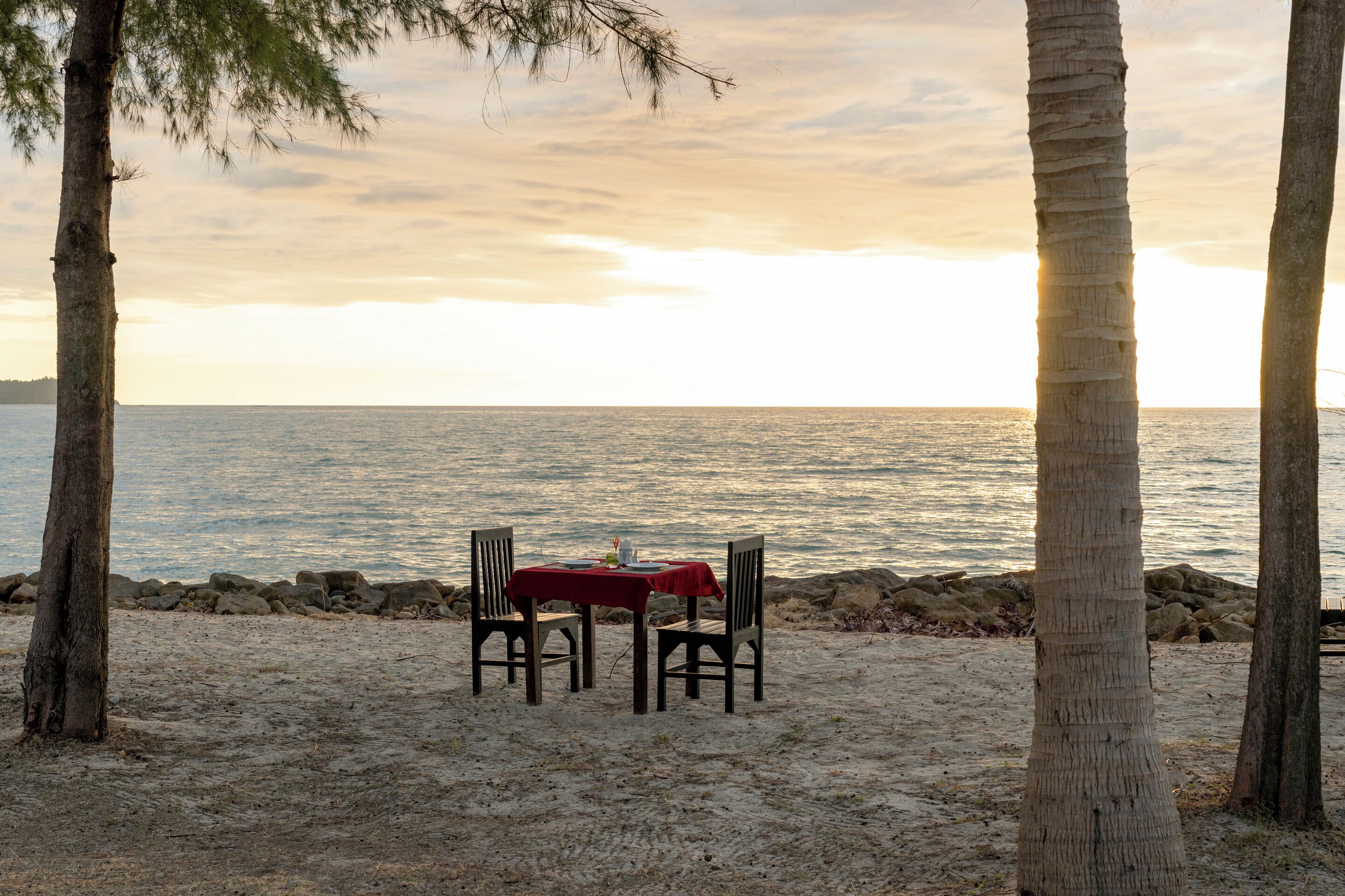 A beachside table with chairs and a red tablecloth, set against a backdrop of the sea and a clear sky.