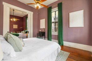 Une chambre à coucher comprenant un lit, un bureau, une chaise, un ventilateur de plafond, une fenêtre avec des rideaux verts et un tableau encadré accroché au mur.
