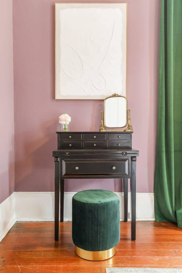 Une pièce comprenant une coiffeuse en bois foncé, un tabouret recouvert d’un tissu vert, une œuvre d’art encadrée et un miroir placé sur la coiffeuse.