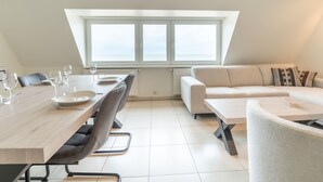 A modern loft apartment with a dining area, a sofa, and a large window offering a view. - Koekoeksnest Penthouse at the beach by Interhome (De Haan)