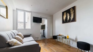 A modern living room with a grey sofa, wooden flooring, a flat-screen TV, a dining area with a small table and chairs, and a wall-mounted artwork.