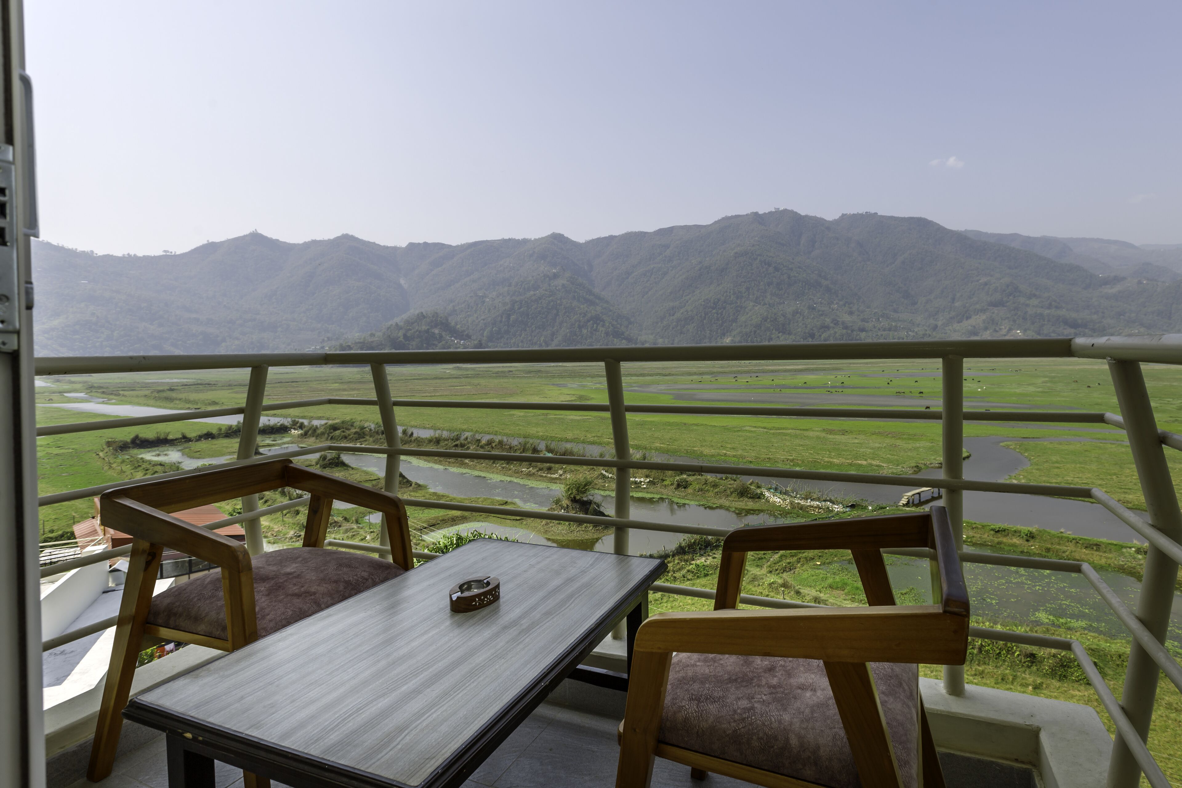 Un balcon avec une table et deux chaises, donnant sur une vallée où l’on aperçoit une rivière et des montagnes.
