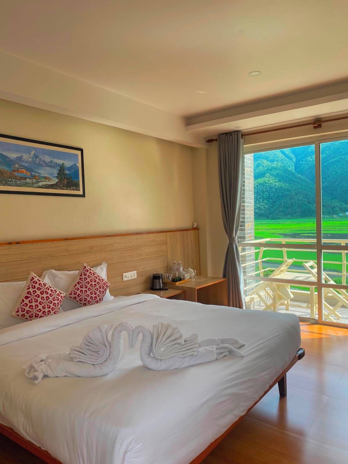 Ein Hotelzimmer mit einem großen Bett, einem Bild an der Wand, Blick auf eine grüne Landschaft durch das Fenster und einem hölzernen Kopfteil.