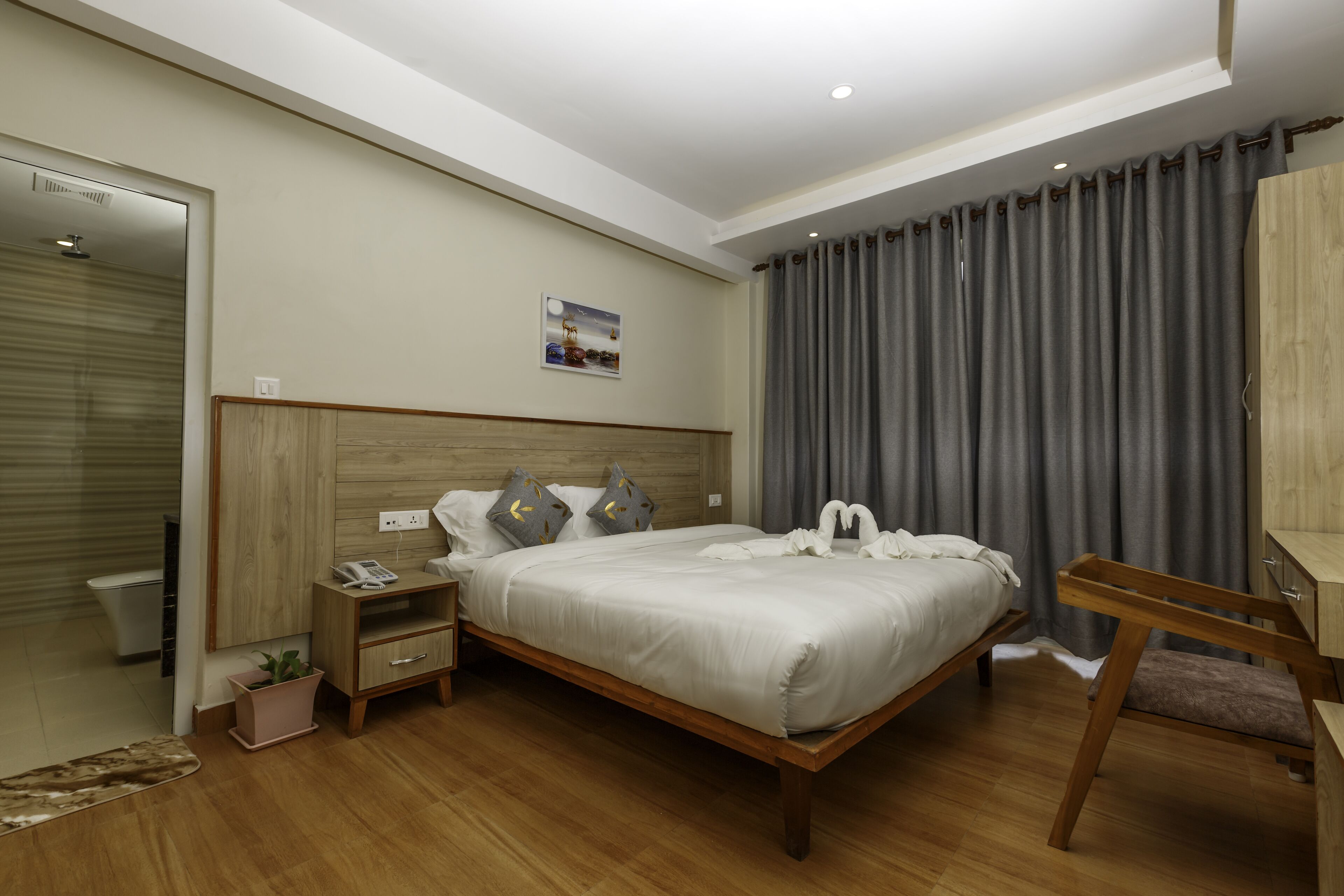 Habitación de hotel moderna con una cama de madera, una mesita de noche con un teléfono, una pequeña planta decorativa y vistas a la ciudad a través de grandes ventanales.