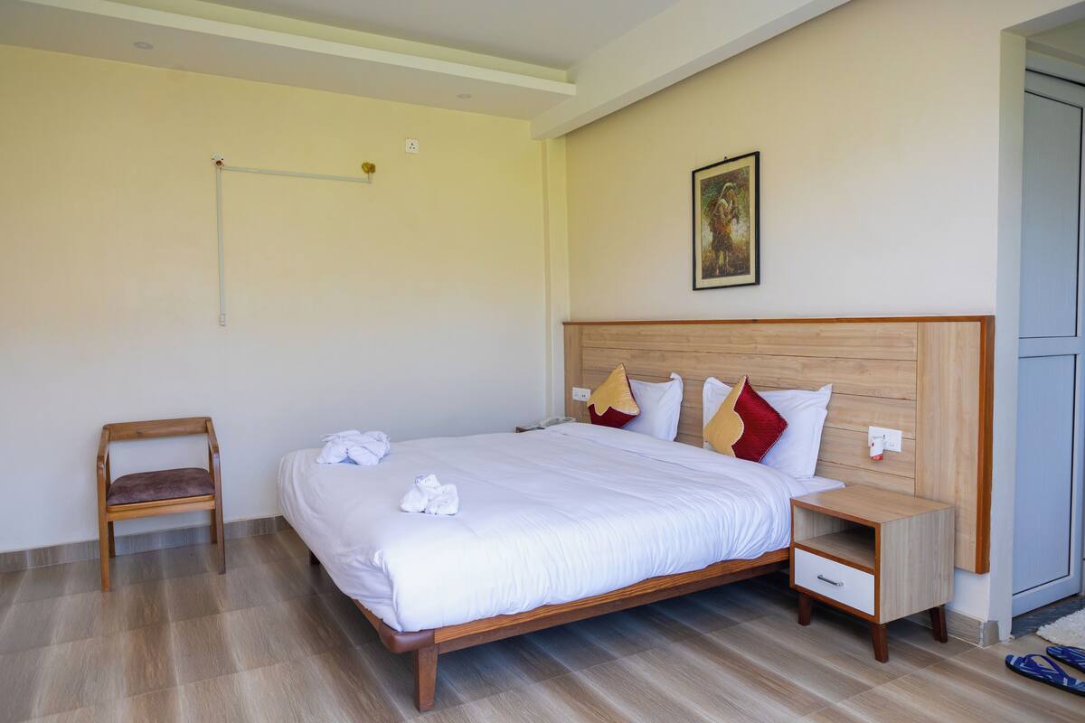 A neatly made bed with white linens, a wooden nightstand with a drawer, a wooden chair, and a painting on the wall.