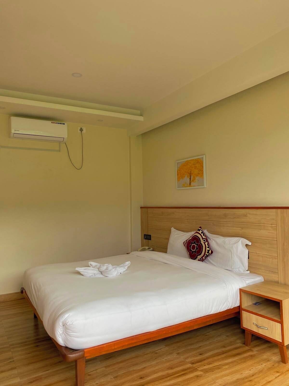 A hotel room with a wooden bed, a nightstand, a wall-mounted air conditioner, and a painting on the wall.