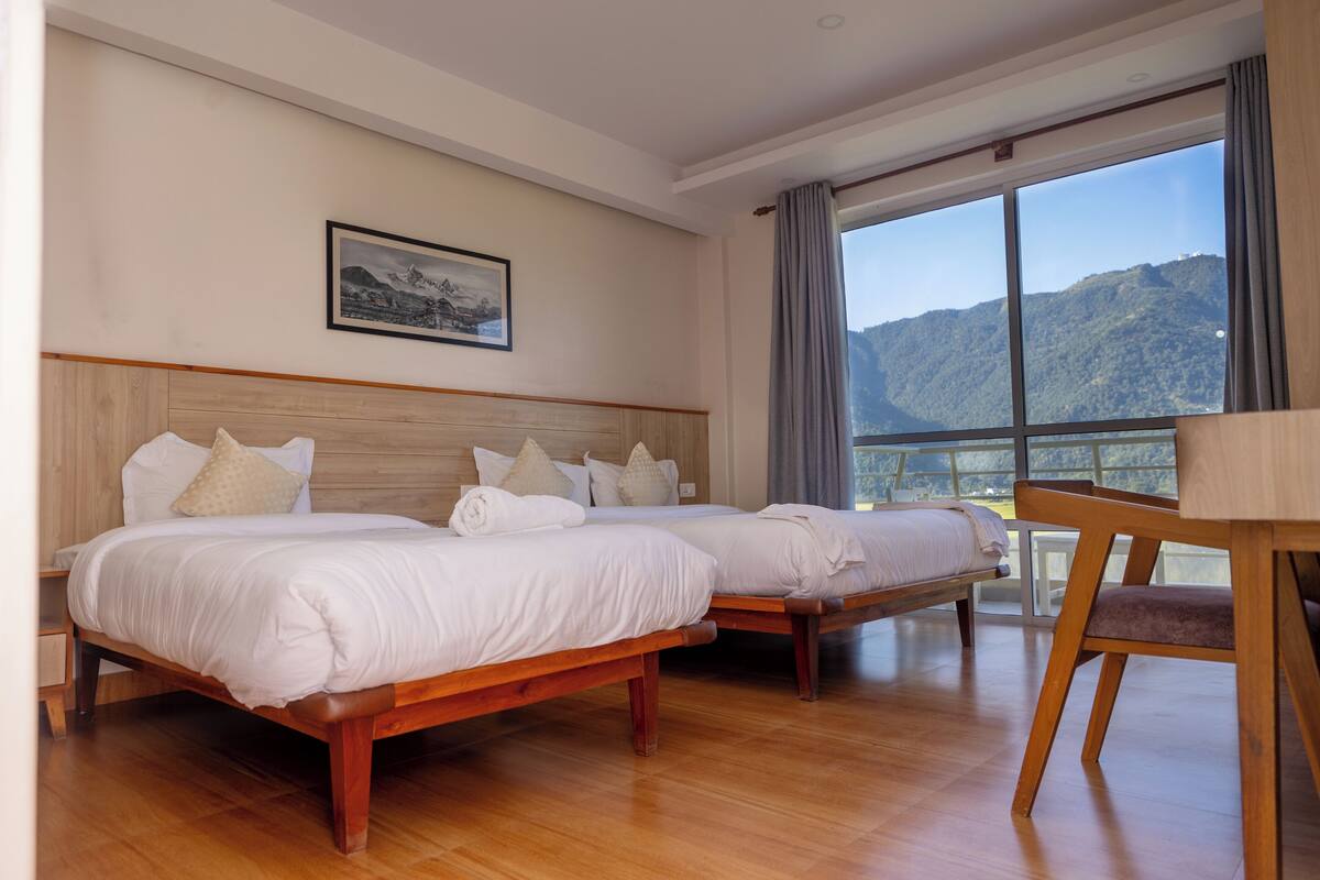 A hotel room with two beds, a wooden headboard, a large window with a mountain view, and a table with a chair.