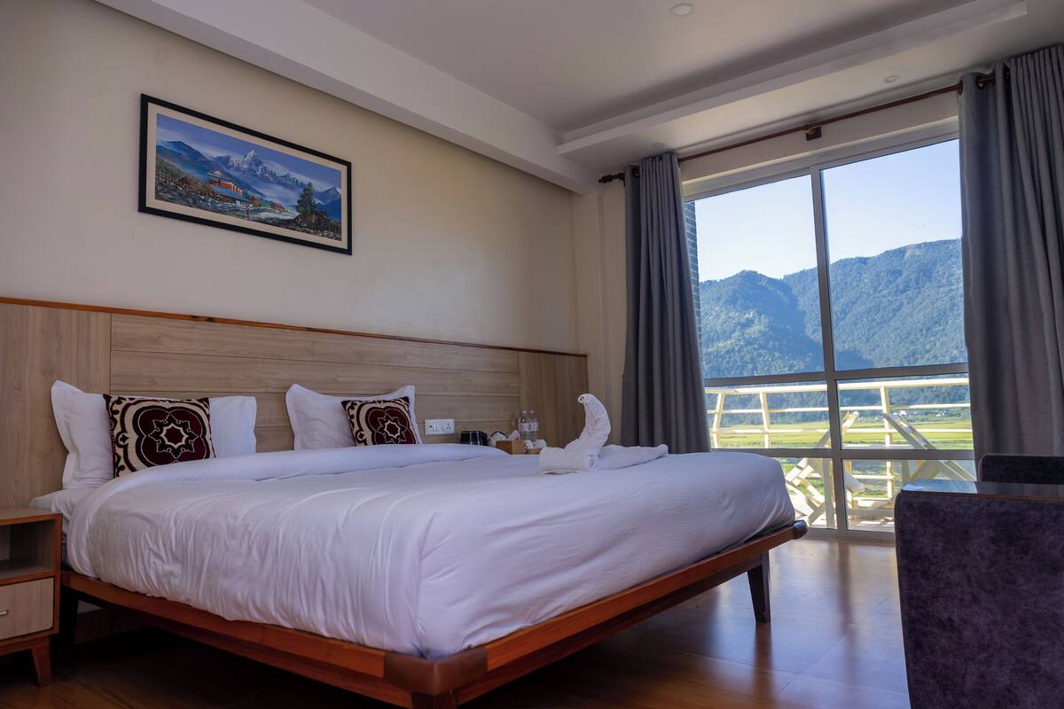 A large bed with white linens, a wooden headboard, and a painting of a mountain landscape on the wall.