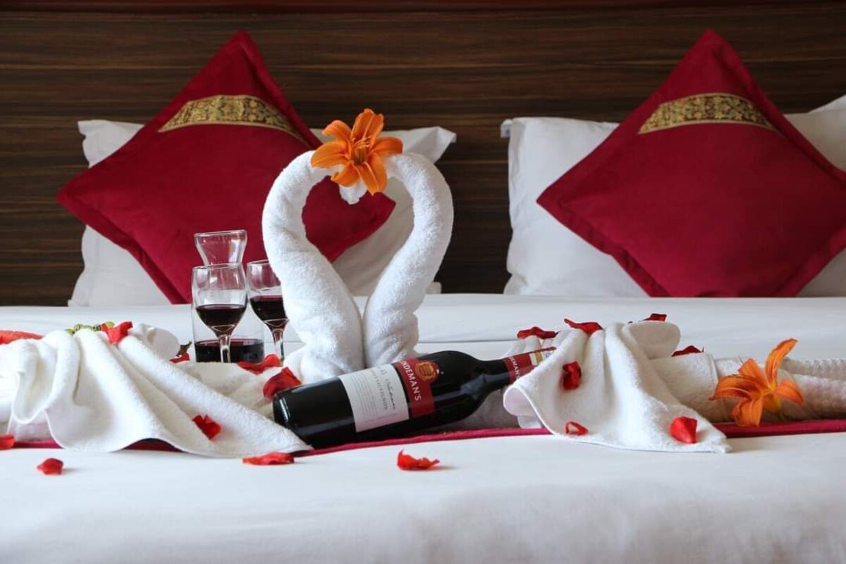 Un lit bien fait, agrémenté d’un bouquet de roses rouges, d’une bouteille de vin et de verres de vin rouge.