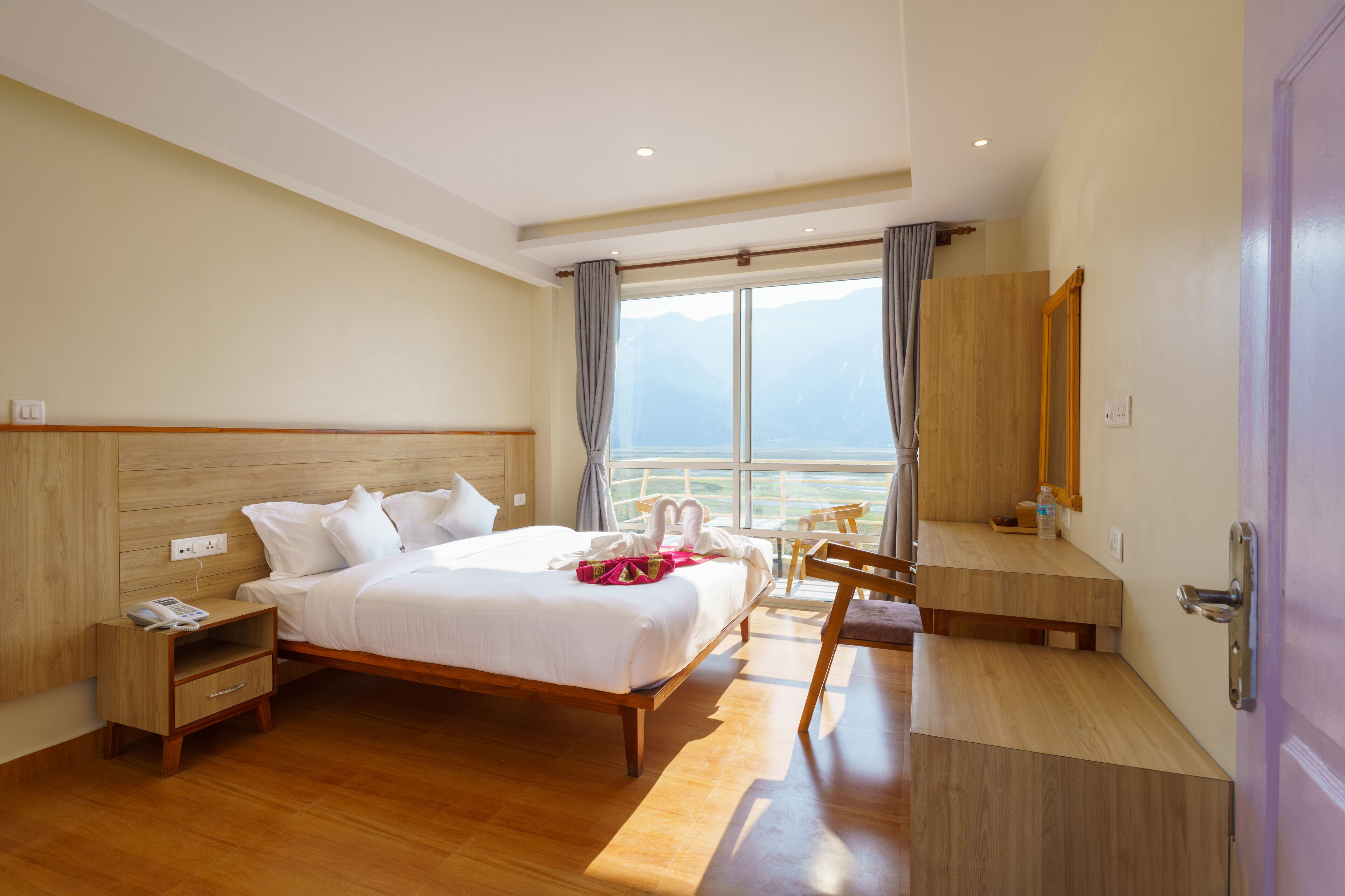 A hotel room with a large bed, wooden headboard, a bedside table with a phone, a chair, a desk, and a large window with curtains.