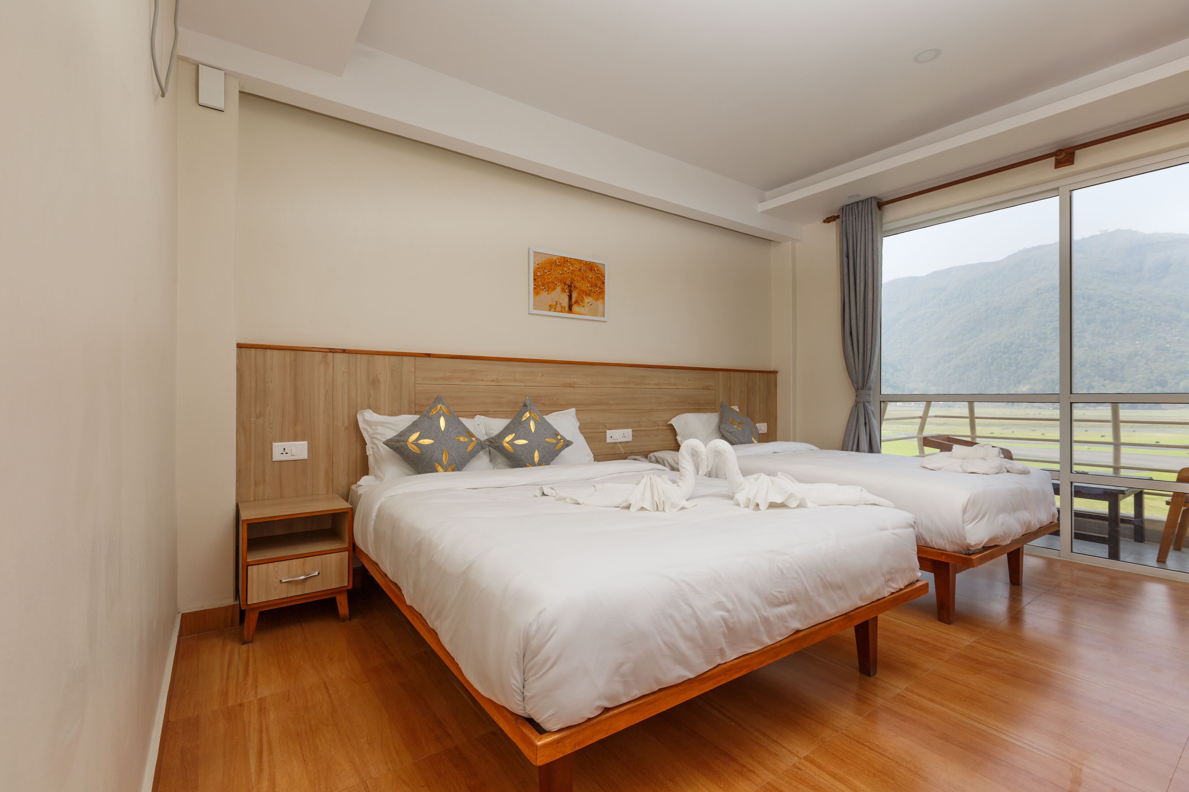 A hotel room with two beds, wooden headboards, a large window with a mountain view, and a small table by the window.