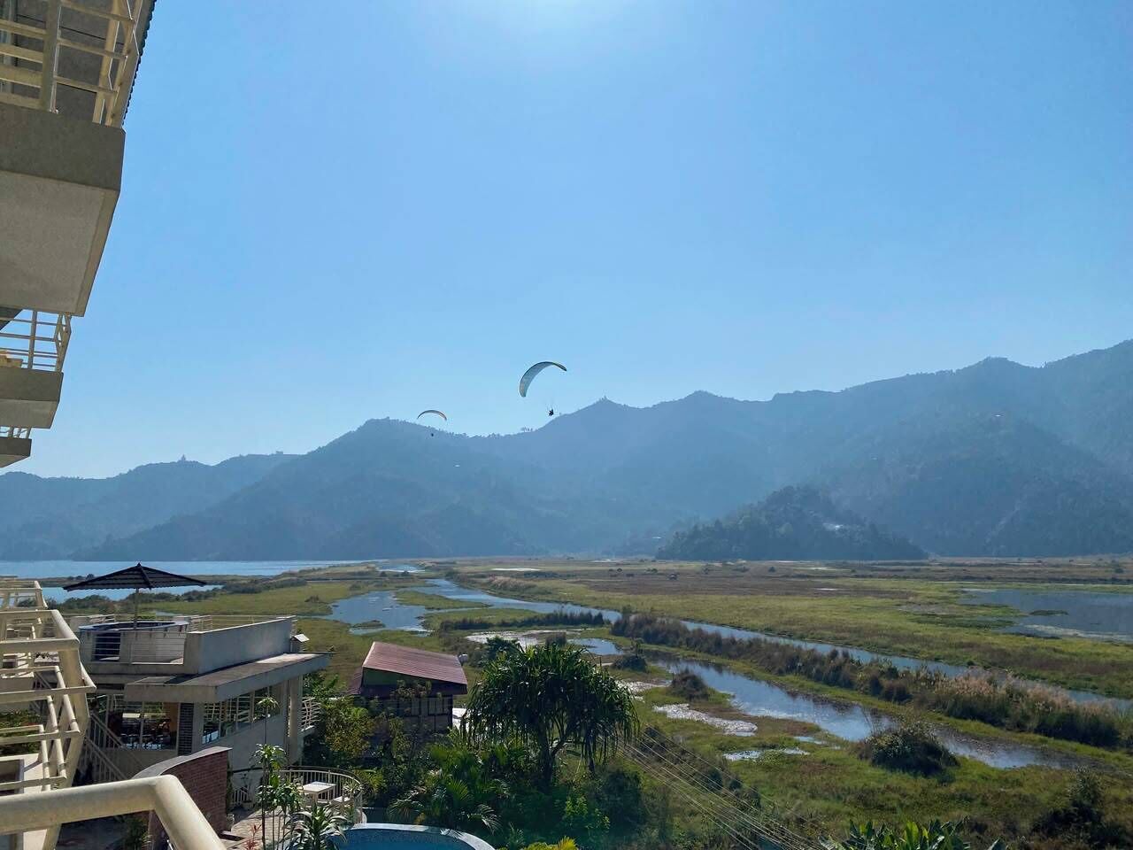 A view of a riverside area with mountains in the background, a building with a balcony, and a person paragliding.