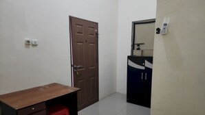 A small, clean room with a wooden desk, a red stool, a brown door, and a white wall with a phone mounted on it. - OYO 93865 Dv Homestay Syariah Kupang (Surabaya)