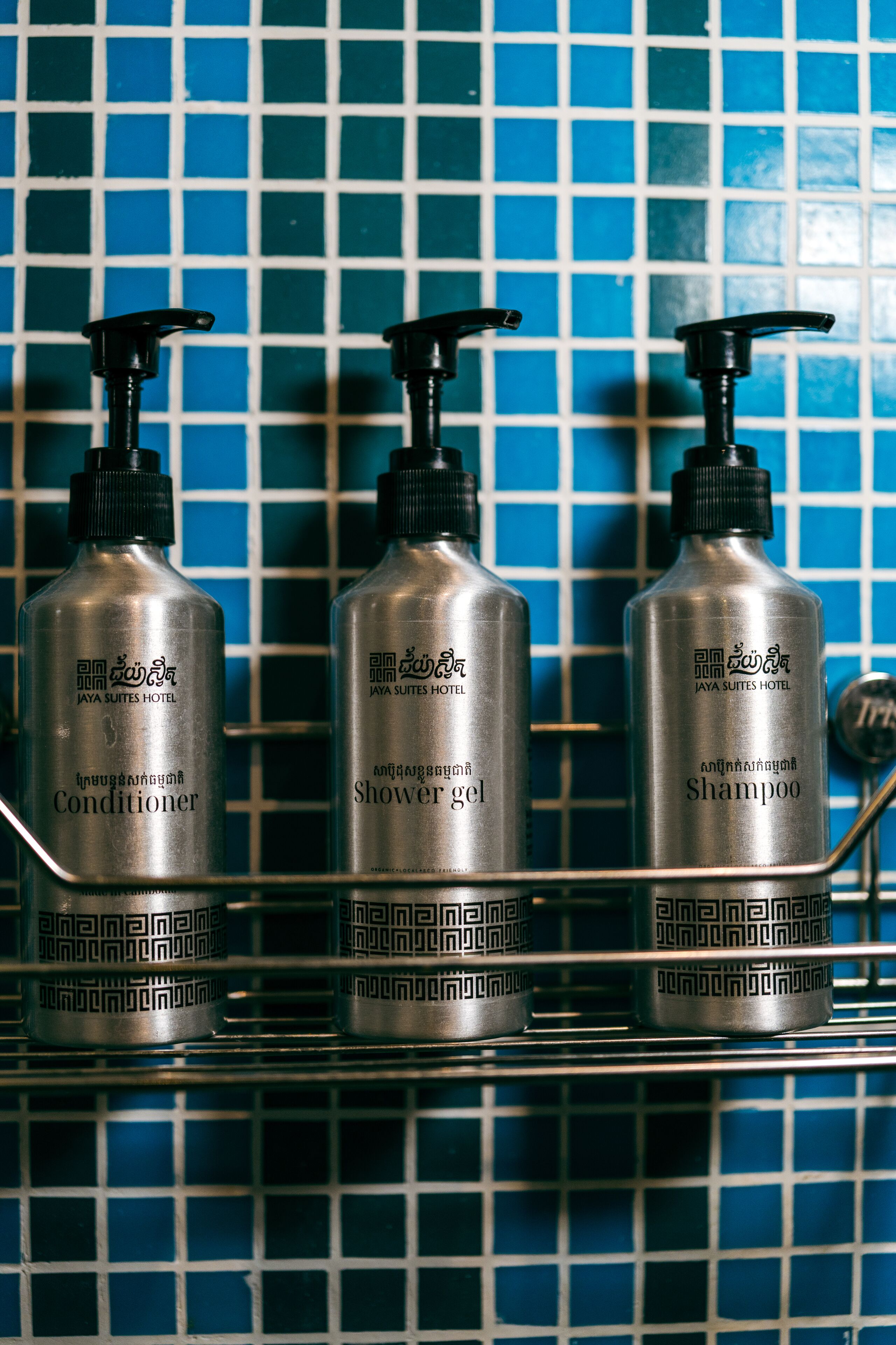 Tres botellas de artículos de aseo del hotel Jaya Suites sobre un estante de metal frente a una pared de azulejos.