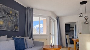 Un salon moderne avec un canapé gris, une table ronde en bois et une vue sur une montagne par la fenêtre.