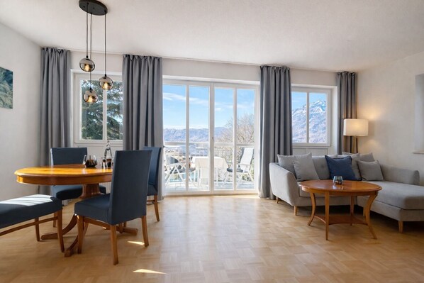 A modern living room with a dining area, large windows, and a view of mountains.