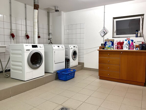 Une buanderie équipée de deux machines à laver à chargement frontal, d’un panier à linge bleu, d’un meuble en bois contenant des produits de nettoyage et d’un sol carrelé.