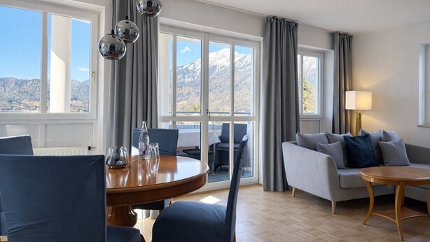 Un salon moderne avec une table à manger en bois, un canapé gris et une petite table ronde. De grandes fenêtres offrent une vue sur les montagnes.