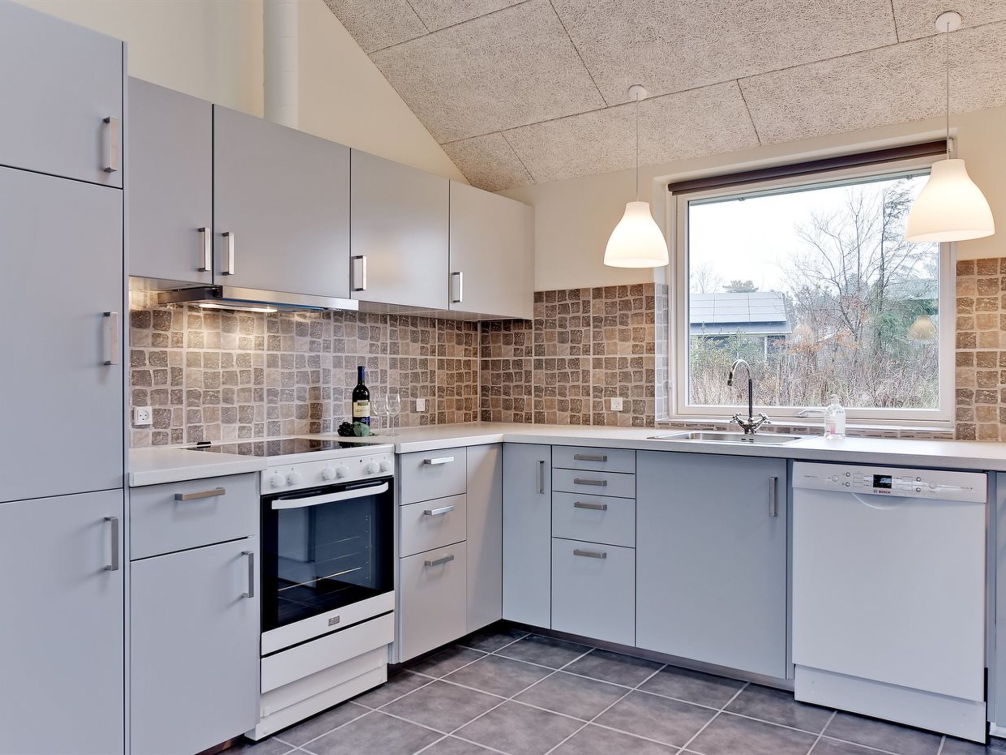 Une cuisine moderne avec des meubles gris, un pare-feu en carrelage, un four encastré et une fenêtre donnant sur les arbres.