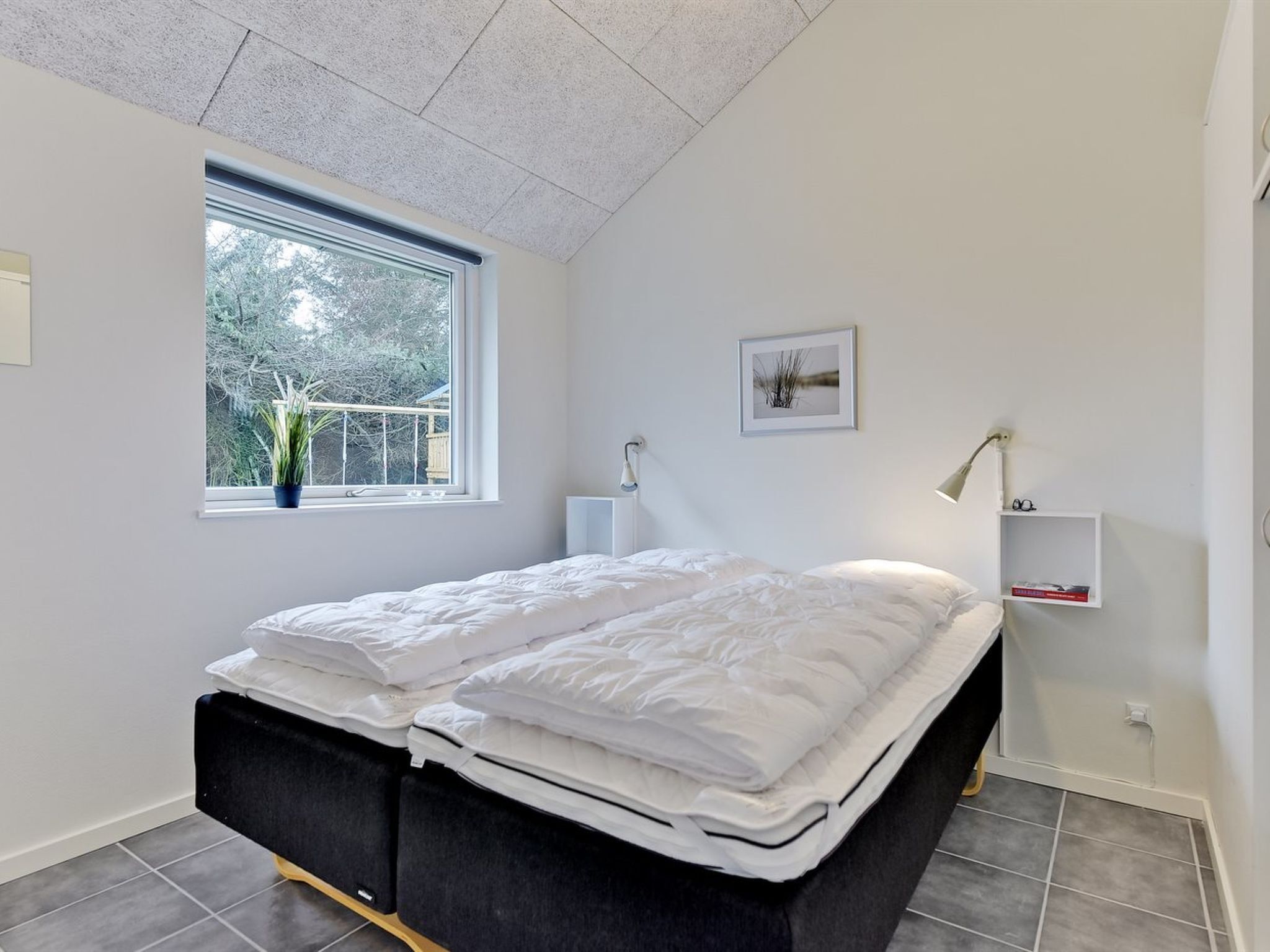 A modern bedroom with a bed, a window, a bedside table, a lamp, and a framed picture on the wall.