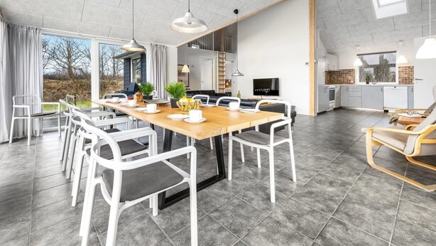 A spacious dining area with a long wooden table and white chairs, a kitchen with modern appliances, and a large window offering a view of the outdoors. - "Talida" - 12km from the sea by Interhome (Oksbøl)