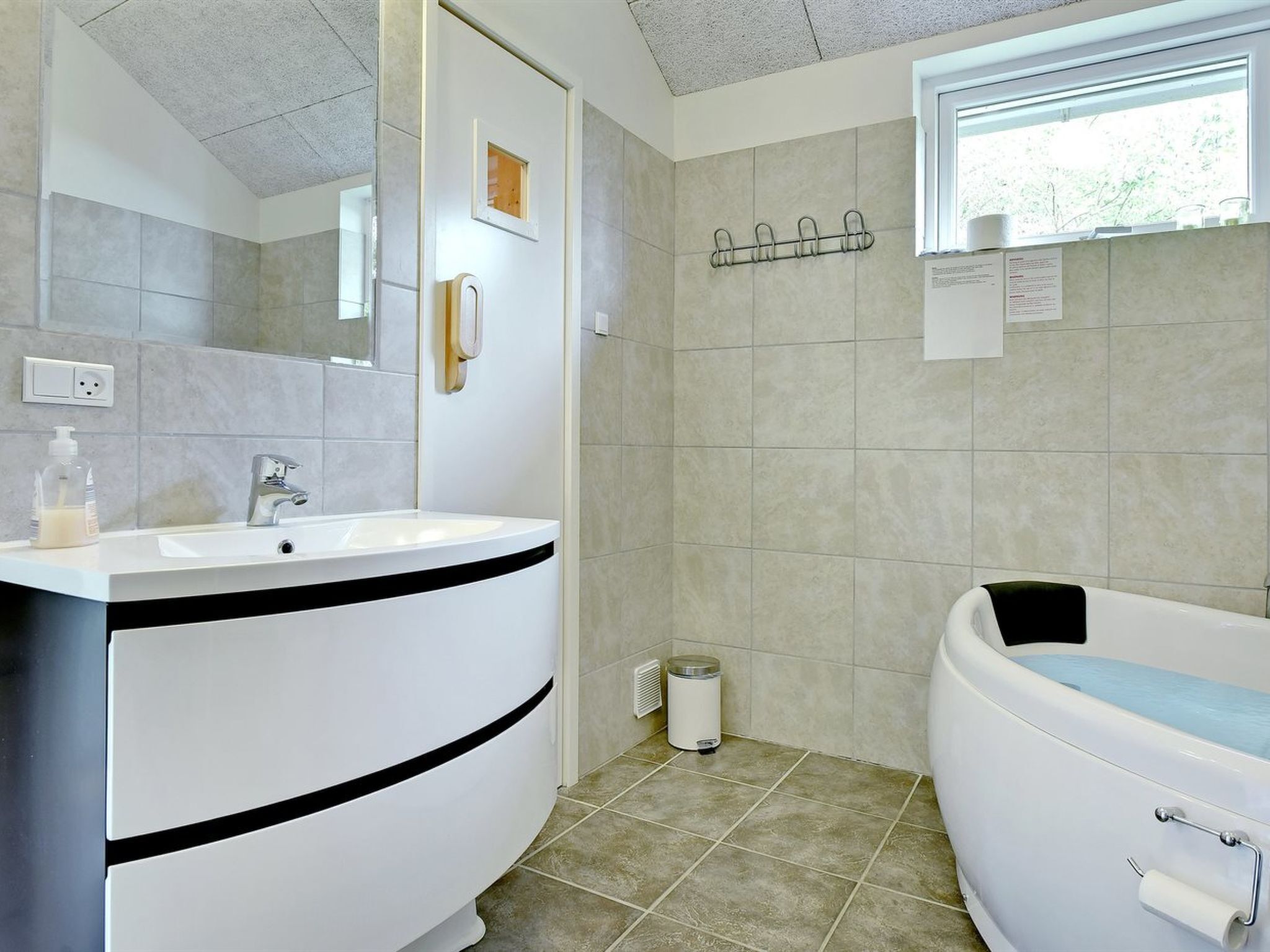 A modern bathroom with a white sink, a large mirror, a bathtub, and a window providing natural light.