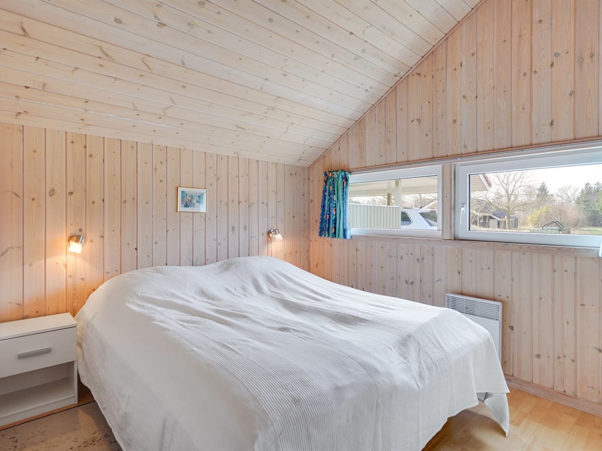 A bedroom with a large bed, wooden walls, a window with a view, and a small bedside table.