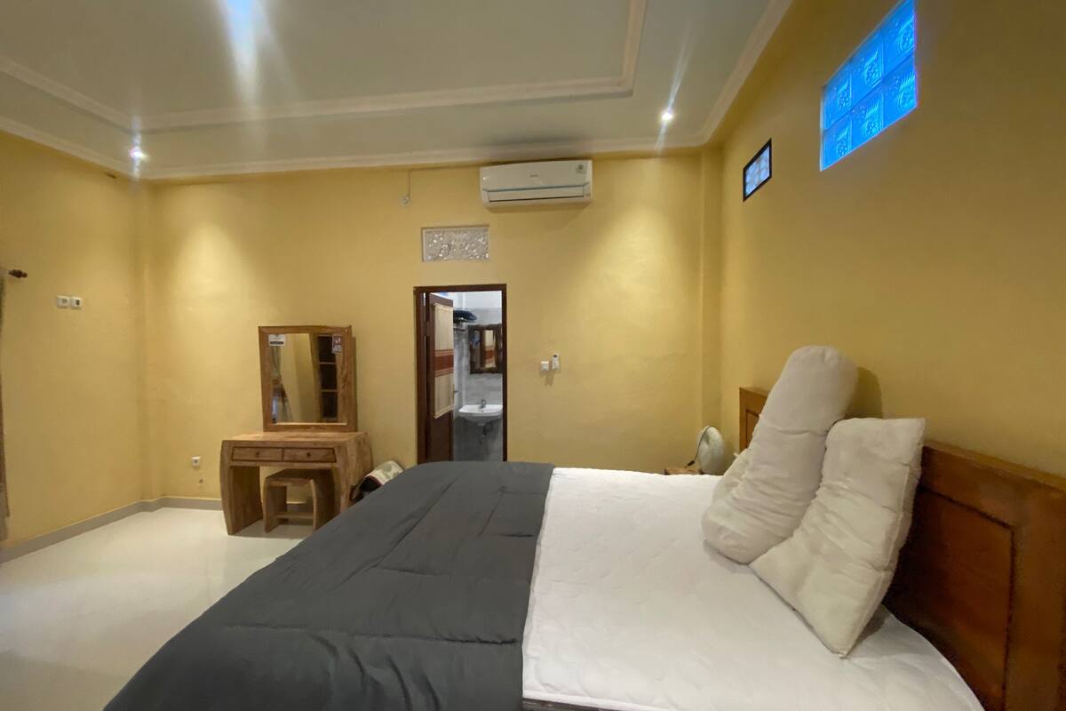 Une chambre d’hôtel avec un lit, un bureau en bois, un miroir, un appareil de climatisation et une porte de salle de bain.