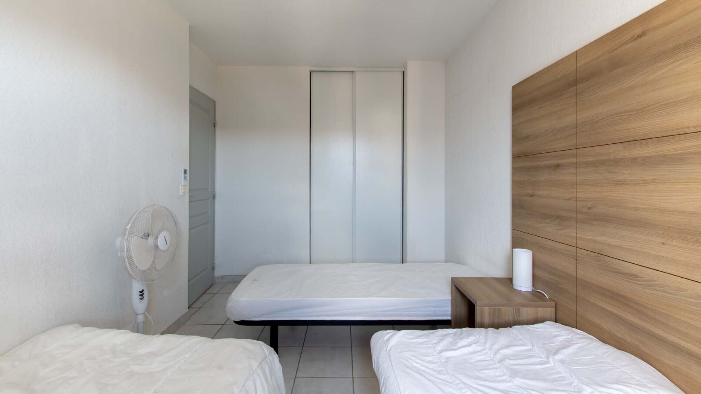 A small, modern hotel room with two single beds, a wooden headboard, a bedside table with a lamp, and a wall-mounted fan.