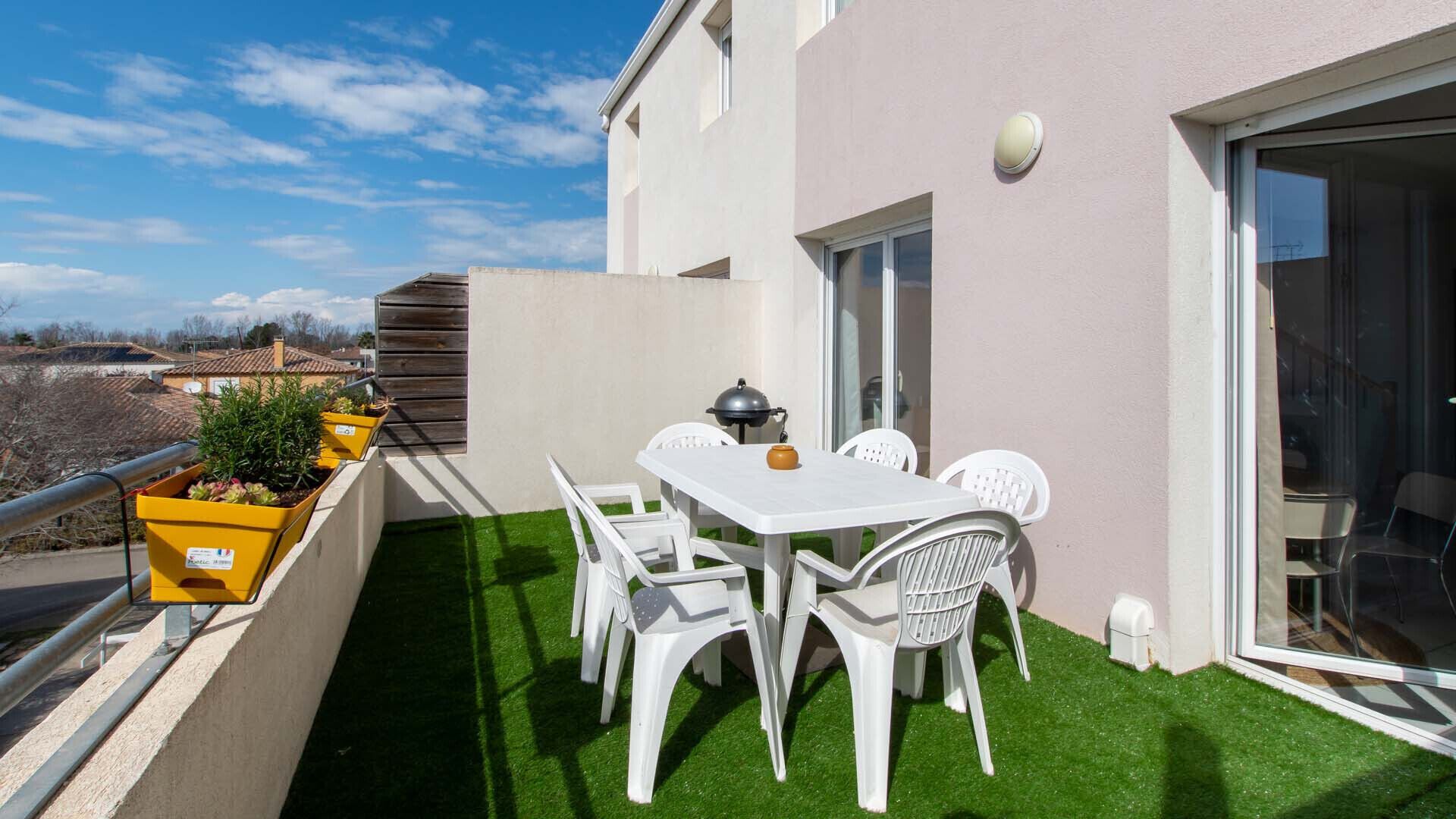 Un balcón con mesa y sillas blancas, una parrilla y plantas en macetas.
